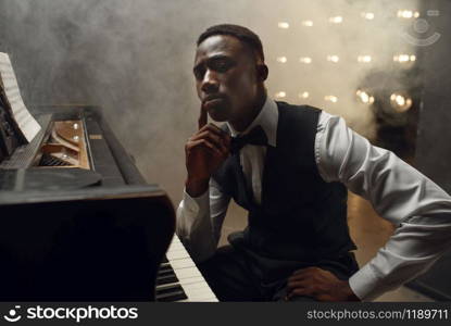 Ebony grand piano musician poses on the stage with spotlights on background. Negro pianist poses at musical instrument before concert. Ebony grand piano musician poses on the stage