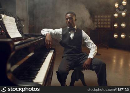 Ebony grand piano player, jazz performer on the stage with spotlights on background. Negro musician poses at musical instrument before concert. Ebony grand piano player, jazz performer