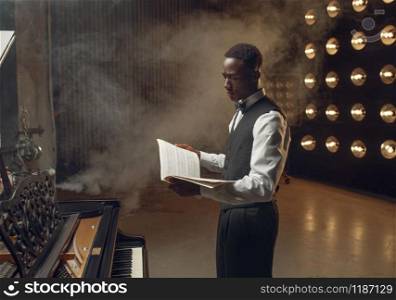 Ebony pianist with music notebook in his hands on the stage with spotlights on background. Negro performer poses at musical instrument before concert