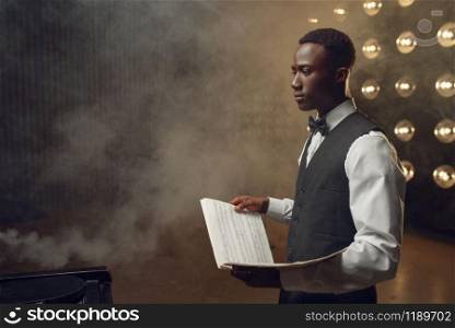 Ebony pianist with music notebook in his hands on the stage with spotlights on background. Negro performer poses at musical instrument before concert. Ebony pianist with music notebook on the stage