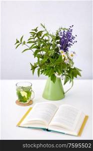 eco and organic concept - cup of green herbal tea, book and flowers in jug on table. herbal tea, book and flowers in jug on table