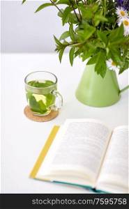 eco and organic concept - cup of green herbal tea, book and flowers in jug on table. herbal tea, book and flowers in jug on table