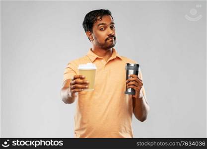 eco living and sustainability concept - young man in polo t-shirt comparing thermo cup or tumbler with disposable paper coffee cup over grey background. man comparing thermo cup or tumbler and coffee cup