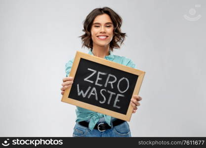 eco living, environment and sustainability concept - portrait of happy smiling young woman in turquoise shirt holding chalkboard with zero waste words grey background. happy woman with chalkboard with zero waste words