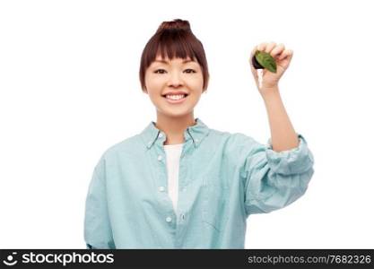 eco living, environment and sustainability concept - portrait of happy smiling young asian woman in turquoise shirt holding car key with green leaf over white background. happy asian woman holding car key with green leaf