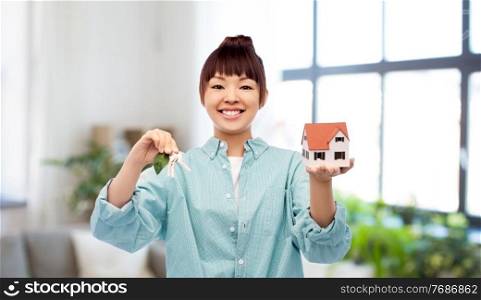 eco living, environment and sustainability concept - portrait of happy smiling young asian woman in turquoise shirt holding toy house and keys with green leaf over home background. smiling asian woman holding house model and keys