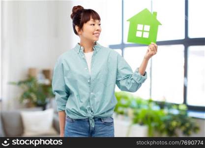 eco living, environment and sustainability concept - portrait of happy smiling young asian woman in turquoise shirt holding green house over home room background. smiling asian woman holding green house