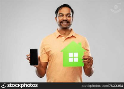 eco living, environment and sustainability concept - smiling young indian man in polo t-shirt holding green house icon and smartphone over grey background. happy indian man with green house and smartphone