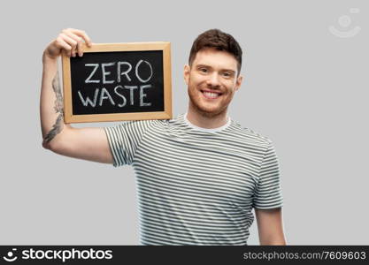 eco living, environment and sustainability concept - smiling young man in striped t-shirt holding chalkboard with zero waste words grey background. smiling man holding chalkboard with zero waste