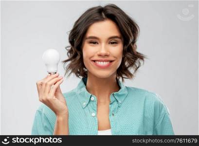 eco living, inspiration and sustainability concept - portrait of happy smiling young woman in turquoise shirt holding energy saving lighting bulb over grey background. smiling woman holding energy saving lighting bulb