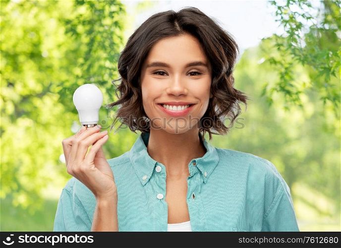 eco living, inspiration and sustainability concept - portrait of happy smiling young woman in turquoise shirt holding energy saving lighting bulb over green natural background. smiling woman holding energy saving lighting bulb
