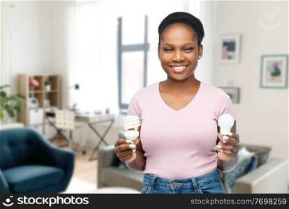 eco living, inspiration and sustainability concept - portrait of happy smiling young african american woman comparing lighting bulbs over home room background. african american woman holding lighting bulbs