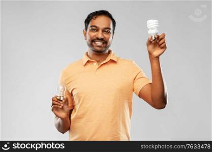 eco living, inspiration and sustainability concept - smiling young indian man in polo t-shirt comparing energy saving light bulb with incandescent lamp over grey background. smiling indian man comparing different light bulbs