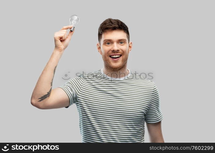 eco living, inspiration and sustainability concept - smiling young man in striped t-shirt holding lighting bulb over grey background. smiling young man holding lighting bulb