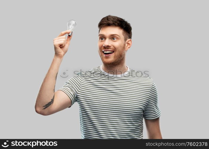 eco living, inspiration and sustainability concept - smiling young man in striped t-shirt holding lighting bulb over grey background. smiling young man holding lighting bulb