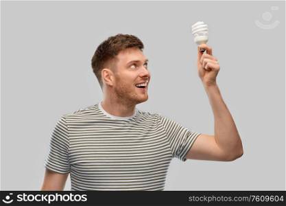 eco living, inspiration and sustainability concept - smiling young man in striped t-shirt holding lighting bulb over grey background. smiling young man holding lighting bulb