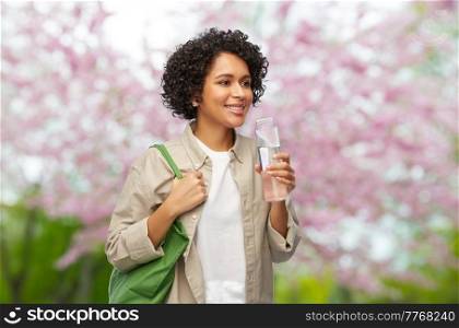 eco living, zero waste and sustainability concept - happy smiling woman with water in reusable glass bottle and shopping bag over blooming tree in spring garden background. happy woman with water in glass bottle and shopper