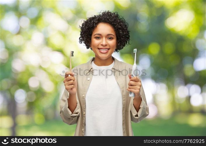 eco living, zero waste and sustainability concept - portrait of happy smiling young woman comparing wooden and electric toothbrushes over green natural background. woman comparing wooden and electric toothbrushes