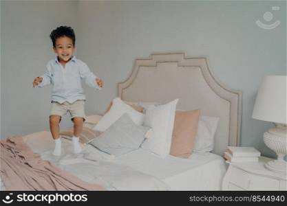 Ecxited mixed race ethnicity kid boy in casual clothes having fun and jumping on big bed, with white and pink beddings and pillows. Photo taken in air movition with child playing indoors. Happy carefree mixed race ethnicity kid boy in casual clothes having fun and jumping on bed