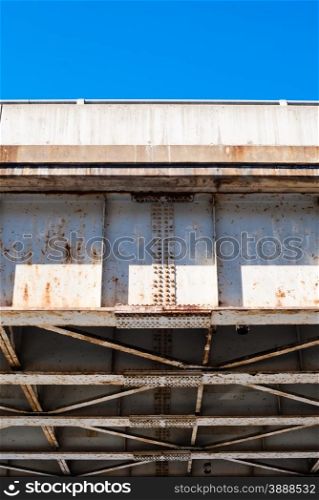 Edge of underside of old rusted steel bridge truss and frame, with rusty rivets and plates, against piece of clear blue sky.