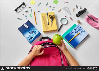 education and learning concept - hands packing pink backpack with green apple, books and school supplies on table. hands with backpack, books and school supplies