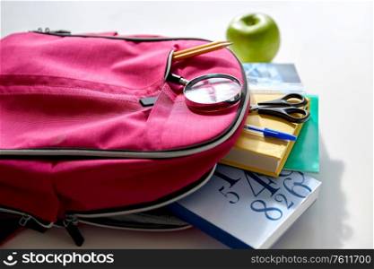 education and learning concept - pink backpack with books and school supplies, green apple on table. backpack with books, school supplies and apple