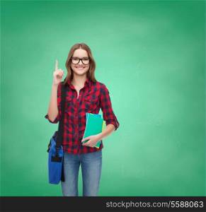 education and people concept - smiling female student in eyeglasses with bag and notebooks showing finger up on green board background