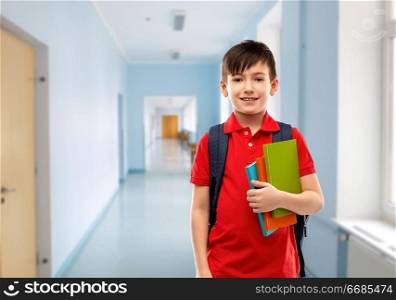 education and people concept - smiling little student boy in red polo t-shirt in glasses with books and bag over school corridor background. smiling student boy with books and school bag