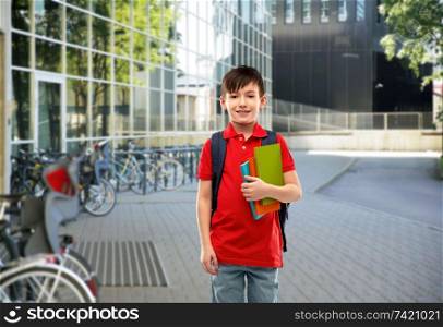 education and people concept - smiling little student boy in red polo t-shirt in glasses with books and bag over school yard background. smiling student boy with books and school bag