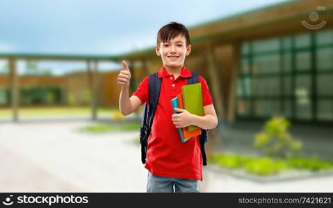 education and people concept - smiling little student boy in red polo t-shirt in glasses with books and bag showing thumbs up over school background. student boy with books and bag showing thumbs up