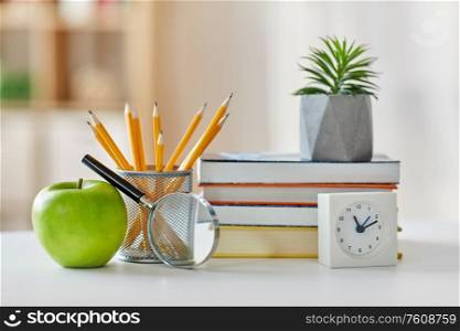 education and school concept - books, magnifier, pencils, plant in flower pot with green apple and alarm clock on table at home. books, magnifier, pencils, apple on table at home