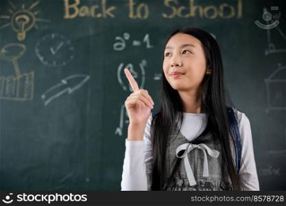 Education Back to school concept. Asian school girl in uniform with backpack on classroom pointing up finger on blackboard, primary child is standing front of class in school thinking of great ideas