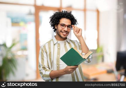 education, business and people concept - happy smiling young man in glasses reading book over office background. happy young man in glasses reading book at office