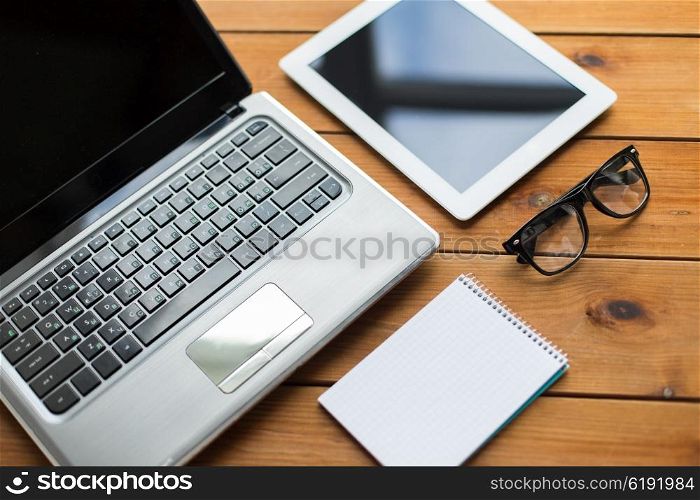 education, business and technology concept - close up of on laptop computer, tablet pc, notebook and eyeglasses on wooden table