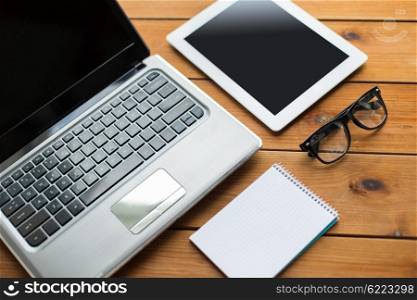 education, business and technology concept - close up of on laptop computer, tablet pc, notebook and eyeglasses on wooden table