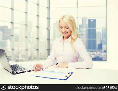 education, business and technology concept - smiling businesswoman with laptop computer and papers sitting over office room with city view window background