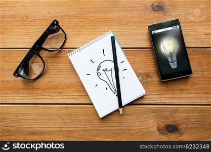 education, business, idea and technology concept - close up of light bulb drawing in notepad with pencil, smartphone and eyeglasses on wooden table