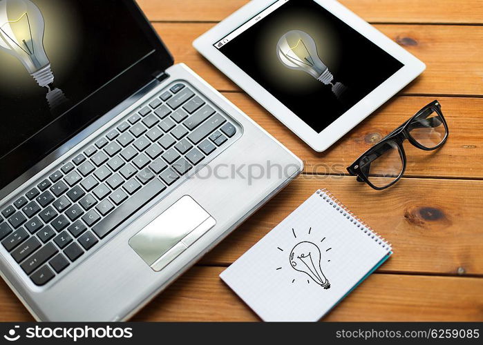 education, business, startup and technology concept - close up of laptop computer, tablet pc and notebook with eyeglasses and light bulbs on wooden table