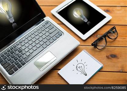 education, business, startup and technology concept - close up of laptop computer, tablet pc and notebook with eyeglasses and light bulbs on wooden table