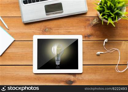 education, business, startup and technology concept - close up of tablet pc computer, laptop and earphones on wooden table with light bulb on screen