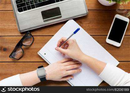 education, business, technology and people concept - close up of woman or student writing to notepad with smart watch, laptop computer and smartphone on wooden table