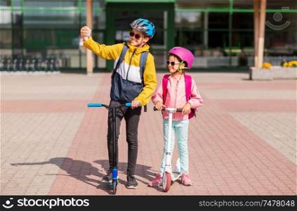 education, childhood and people concept - happy school children in helmets with backpacks riding scooters and taking selfie by smartphone outdoors. happy school kids with scooters taking selfie