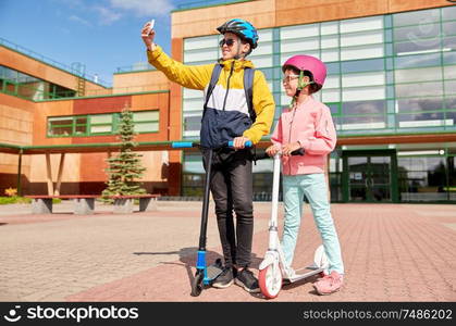 education, childhood and people concept - happy school children in helmets with backpacks and scooters taking selfie by smartphone outdoors. happy school kids with scooters taking selfie