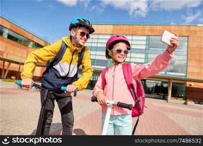 education, childhood and people concept - happy school children in helmets with backpacks and scooters taking selfie by smartphone outdoors. happy school kids with scooters taking selfie
