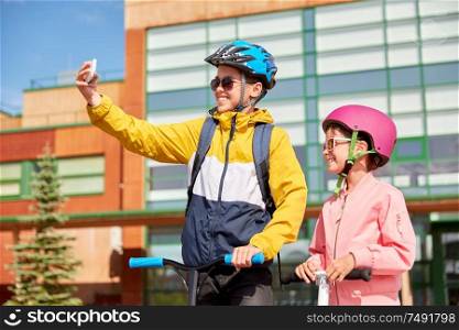 education, childhood and people concept - happy school children in helmets with backpacks and scooters taking selfie by smartphone outdoors. happy school kids with scooters taking selfie