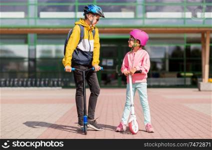 education, childhood and people concept - happy school children in helmets with backpacks riding scooters outdoors. happy school children in helmets riding scooters