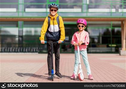 education, childhood and people concept - happy school children in helmets with backpacks riding scooters outdoors. happy school children in helmets riding scooters