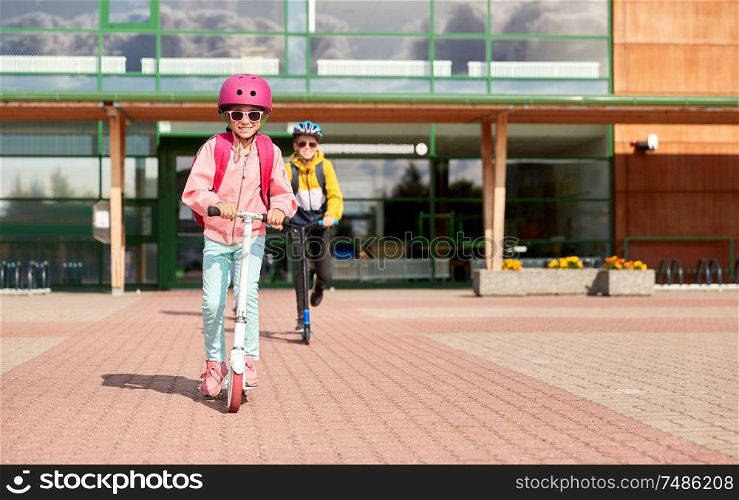 education, childhood and people concept - happy school children in helmets with backpacks riding scooters outdoors. happy school children riding scooters outdoors