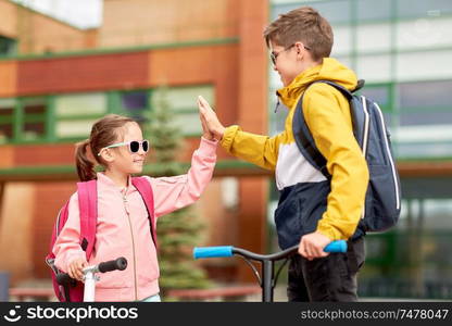 education, childhood and people concept - happy school children with backpacks and scooters making high five outdoors. happy school children with backpacks and scooters