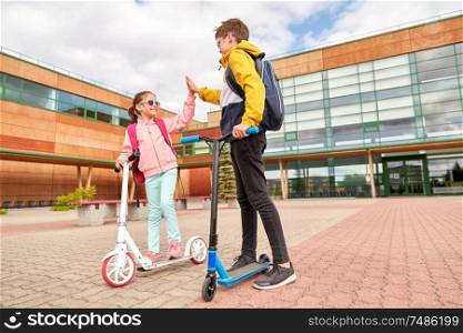 education, childhood and people concept - happy school children with backpacks and scooters making high five outdoors. happy school children with backpacks and scooters
